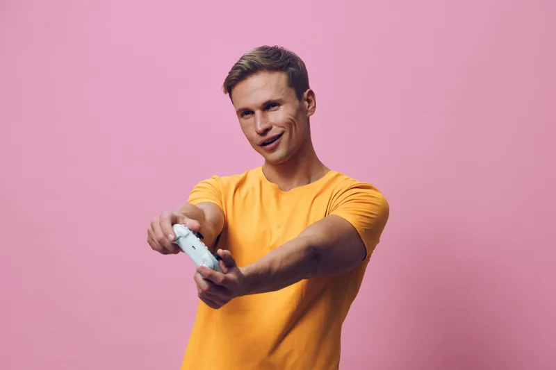 Young smiling man play video game hold console controller wear orange t-shirt pink background focus enjoyment concept digital detox relaxation technology
