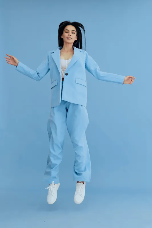 young woman jumping wearing light blue suit with white sneakers and lace top on blue background, cheerful expression in stylish fashion outfit