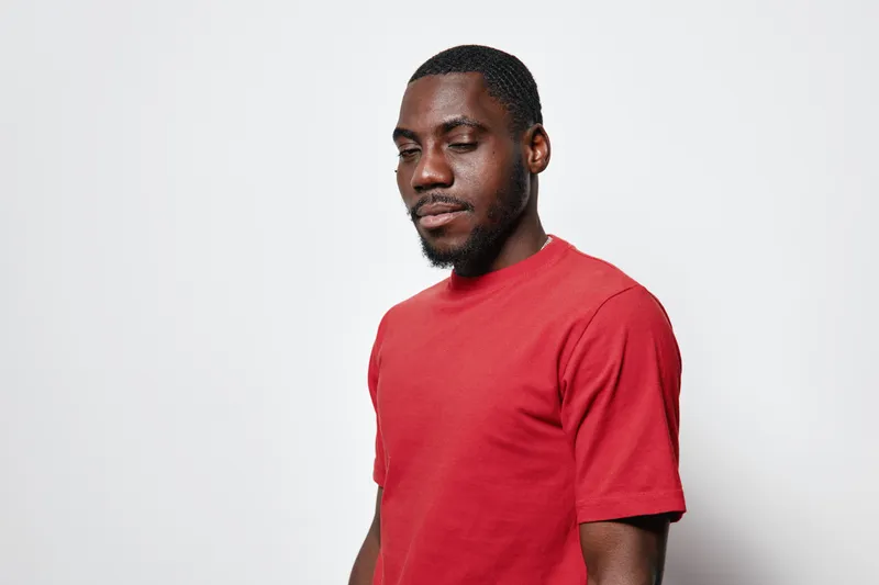 African American man with dark skin wearing red t shirt standing against white background with eyes closed and calm expression in studio portrait. Male model with short hair and beard.