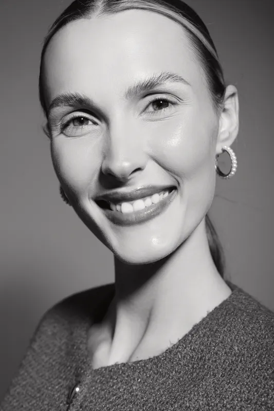 Portrait of smiling Caucasian woman with fair skin and slicked back hair wearing earrings and a textured top. Studio lighting highlights her natural beauty and confident expression.