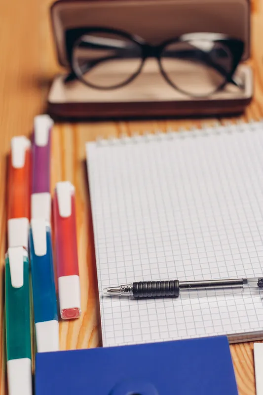 На рабочем столе лежат блокноты, фломастеры, канцелярские принадлежности и очки в футляре