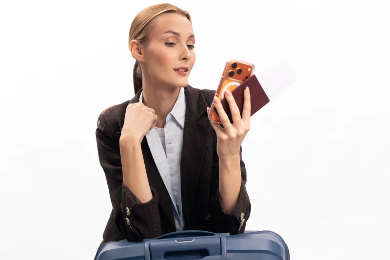 Young woman in casual jacket holding phone and passport looks to side, sitting behind suitcase. Concept of travel planning, waiting, and preparation for trip.