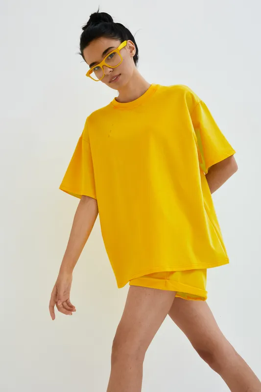 young woman wearing oversized yellow outfit and matching sunglasses posing indoors with white background, casual style, bright fashion look