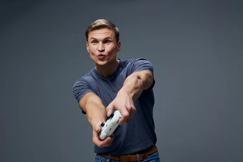 Fun young man making playful face while holding gaming controller pointing forward isolated on gray background studio portrait casual clothing lifestyle concept