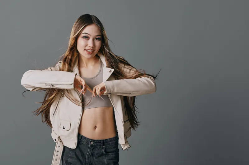 Young woman with long hair wearing light jacket and crop top posing and smiling against gray background. Casual style, confident female model with natural makeup and denim pants.
