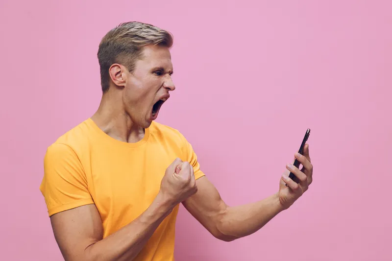 Vibrant young man in yellow t-shirt celebrating success while looking at smartphone screen isolated on pink background. Studio portrait showcasing excitement, triumph, happiness, achievement, expressi