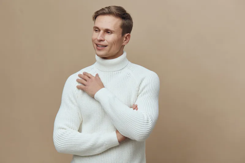 Smiling young man wearing white turtleneck sweater looking away isolated on beige background studio portrait. People lifestyle concept