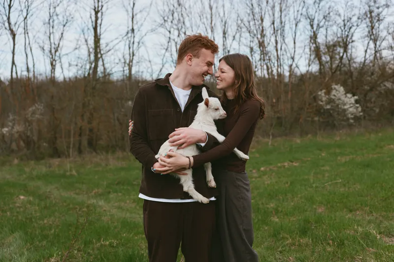 Cheerful couple outdoors, embracing a goat kid, smiling together in a sunlit field, calm countryside mood, natural colors, relaxed posture, tender connection, casual fashion