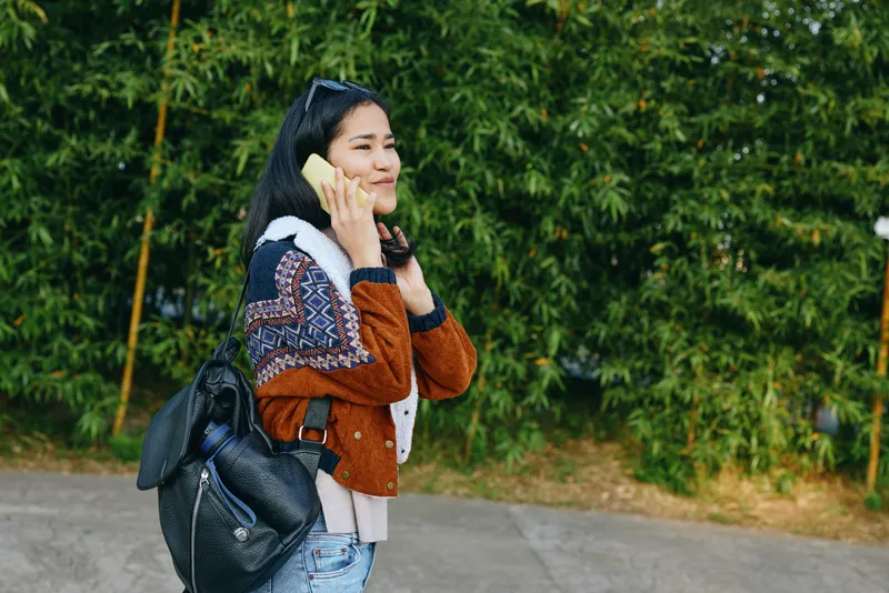 Woman talking on a phone outdoors with a backpack and sweater, strolling along a sidewalk beside a green hedge, casual urban moment, lifestyle and communication