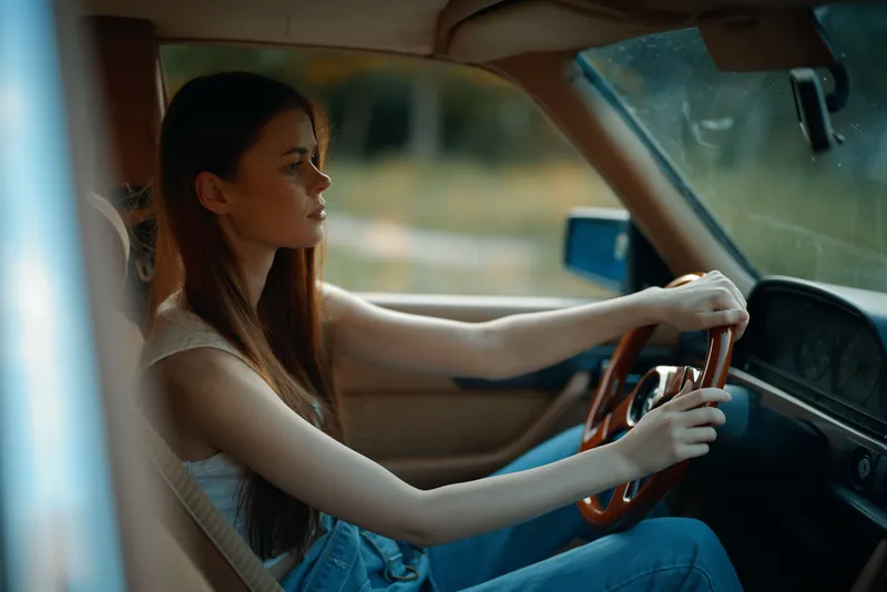 Young woman driving an old car, showcasing confidence and elegance, surrounded by nature, with a focus on her stylish attire and serene ambiance.