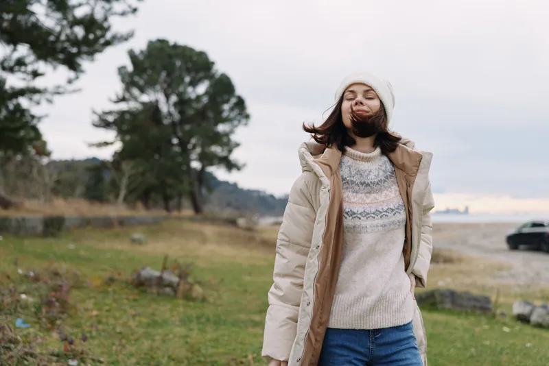 Winter portrait of a woman outdoors wearing a knit sweater, beige coat and beanie, wind blowing her hair, capturing casual fashion and cold countryside mood.