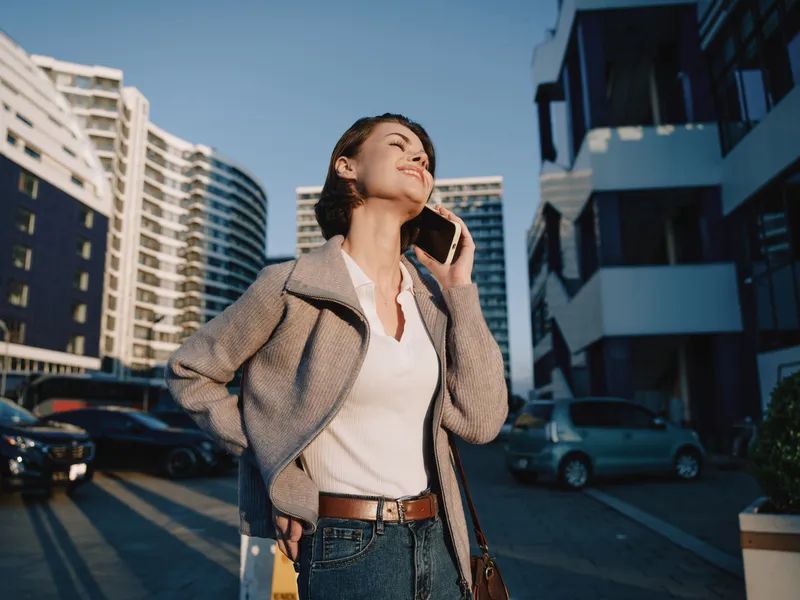 A cheerful business professional stands outdoors in an urban city setting, texting on a smartphone. She wears smart casual attire, smiles warmly, and conveys confident, energetic workplace momentum.