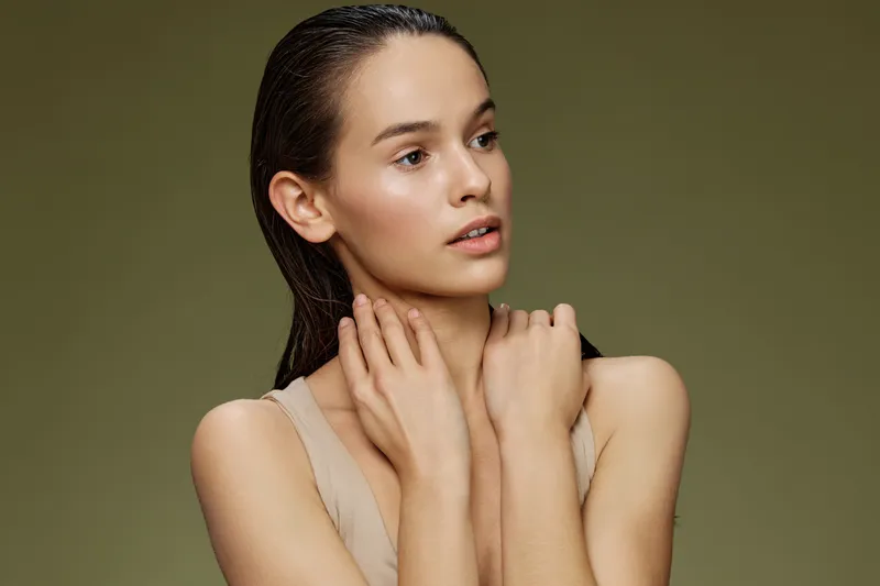 Young woman with wet hair, wearing a nude tank top, posing gracefully against a soft green background, exuding calm and natural beauty