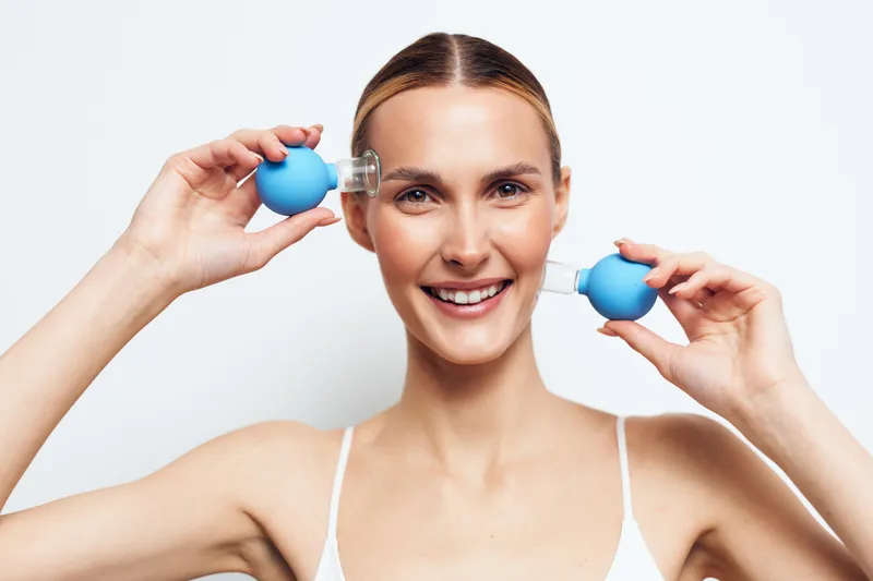 Woman smiling skincare serum spray face beauty self care, young woman holds blue dropper bottles near cheeks, bright studio lighting on light background, concept of skincare wellness