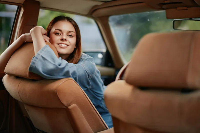 Young woman in denim jacket smiling inside a car, expressing happiness and relaxation, with a soft background and warm color palette.