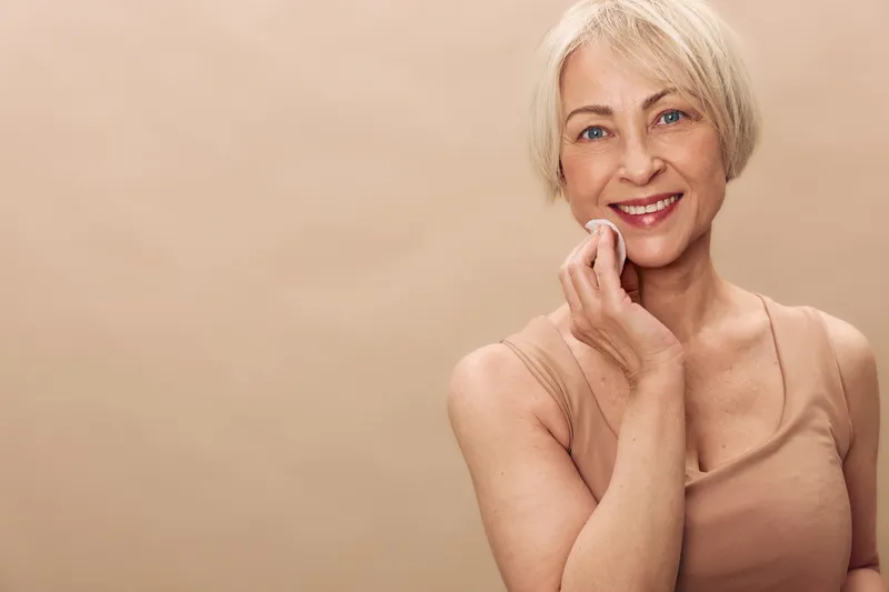 Smiling mature woman with short blonde hair applies facial cotton pad on clean skin against beige background. Concept of skincare, beauty routine, natural aging, and self-care for women.
