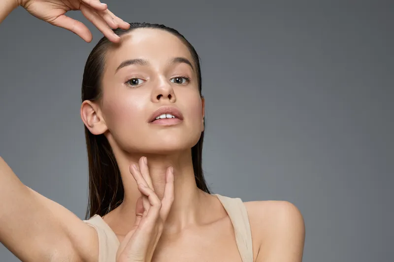 Elegant young model with natural makeup gracefully showcasing her skin, posing against a minimalistic grey background, highlighting beauty and freshness