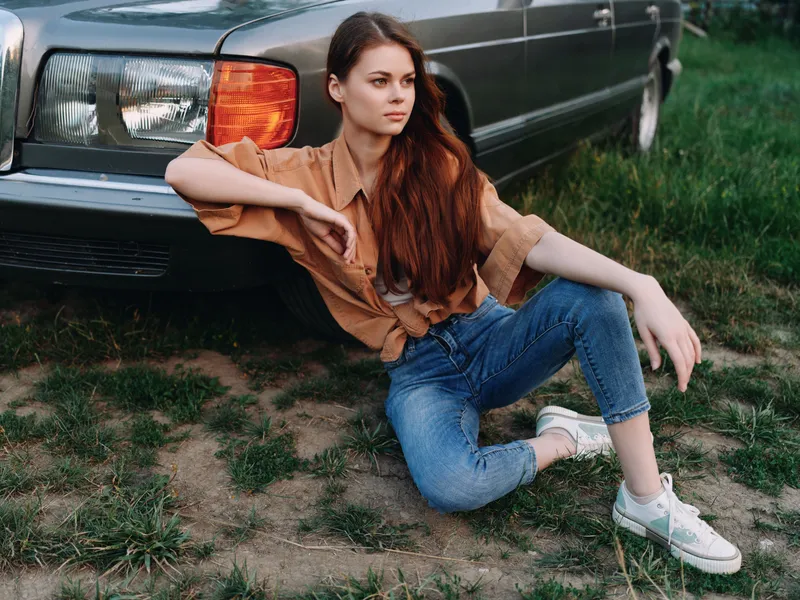 Young woman with long hair sitting casually by a vintage car in denim and a shirt, exuding relaxed confidence in a natural green setting.