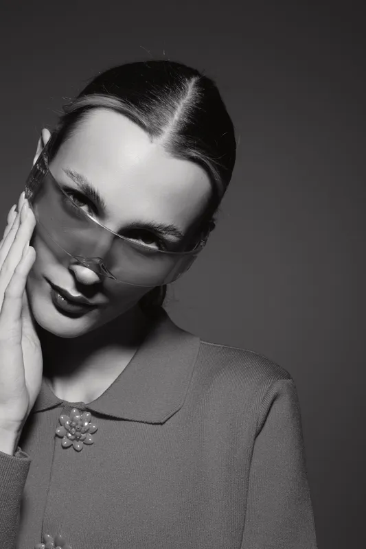 Elegant white woman with light skin wears transparent sunglasses and a textured jacket with a decorative brooch. Studio portrait in black and white with moody lighting and vintage style.