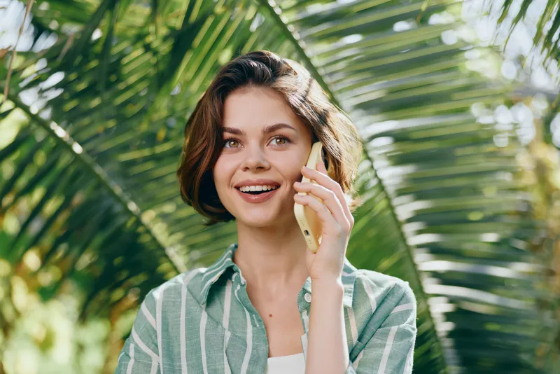 Woman on phone outdoors among lush greenery, smiling with a bright expression, casual attire and natural light highlight her cheerful mood, conveying a moment of connection and ease.
