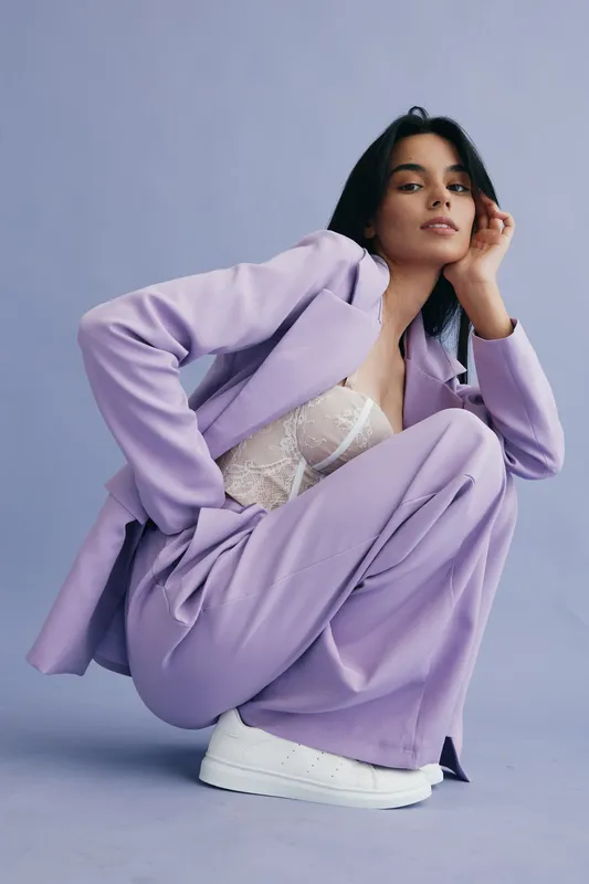 confident woman in lilac pantsuit crouching against pastel purple background wears white sneakers and lace top, showing modern fashion and relaxed pose, stylish young adult