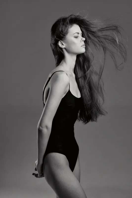 Black and white portrait of a young woman with long hair in a black bodysuit posing sideways with closed eyes.