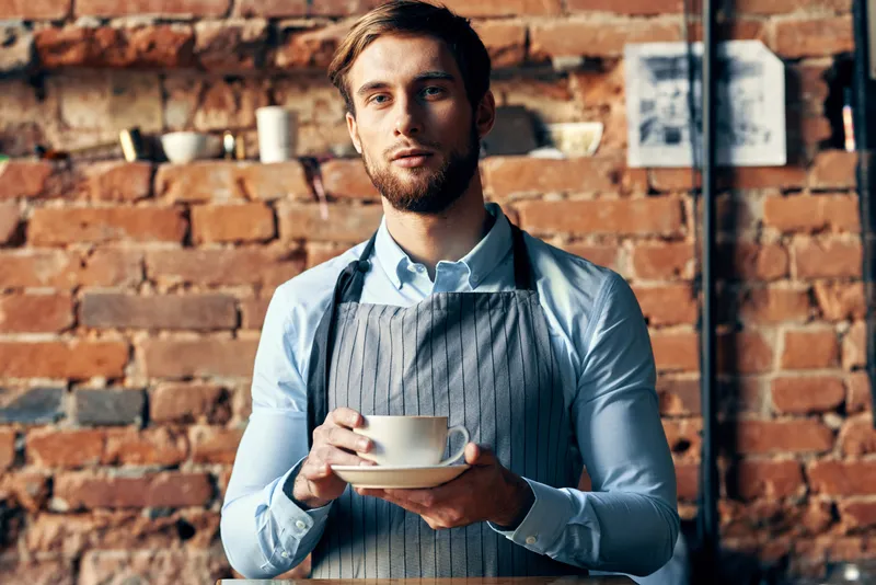 Мужчина в фартуке, бариста, профессионально работает, держа в руке чашку кофе