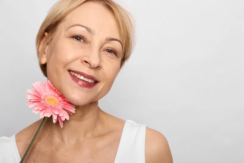 Woman smiling flower closeup blonde portrait skincare wellness beauty studio lighting, short hair, white top, pink blossom on neck, healthy skin, calm mood