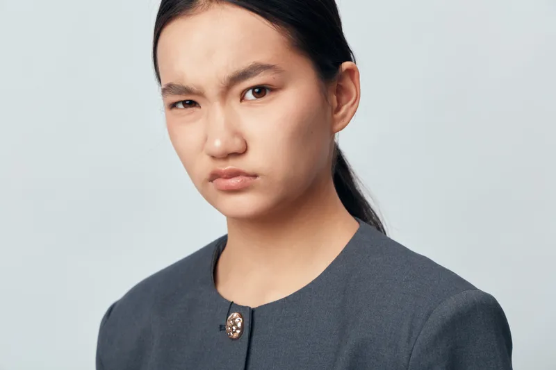 Young woman with dark hair and serious expression looking skeptical or doubtful against plain light background. Portrait with natural skin, dressed in simple dark clothing and neutral mood.