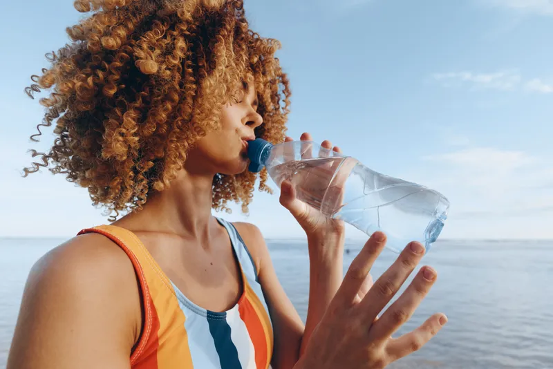 Smiling woman with curly hair drinks water from a bottle by the seaside, wearing a striped sleeveless top. Close-up on a bright blue sky, calm water, and soft sunlight.