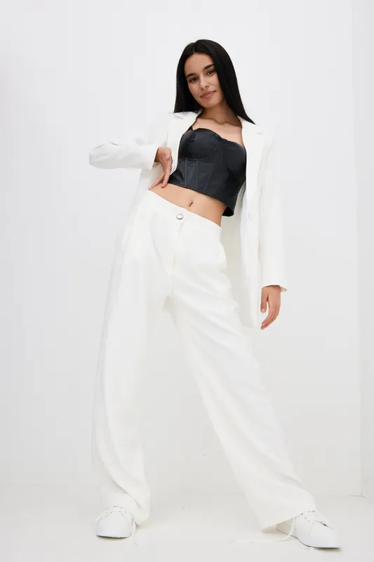 Stylish woman in white wide pants and blazer with black corset top posing confidently against plain white background, showcasing modern fashion and casual elegance.