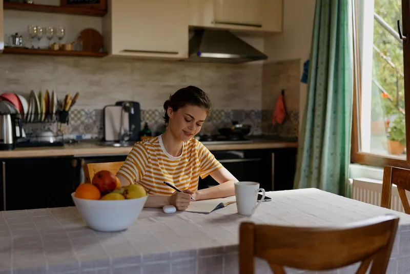 A woman sits at a kitchen table writing in a notebook, bathed in natural light with a fruit bowl and coffee mug nearby, conveying a calm, homey morning mood.