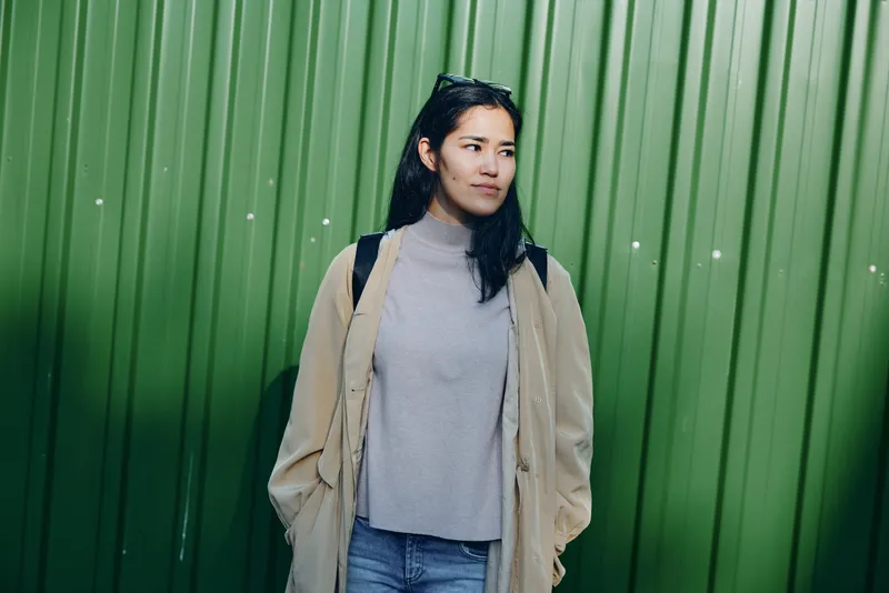 Woman in a casual coat and sweater stands against a vivid green metal wall, urban street fashion, relaxed pose, natural light, candid mood, contemporary lifestyle