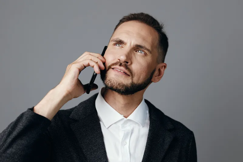 A professional man in a suit speaks on a mobile phone, showing thoughtful emotion against a solid colored background, conveying focus and confident leadership.