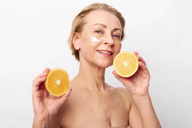 Mature woman with short blonde hair holds two halves of an orange near her face with cream under eyes. Concept of natural skincare, healthy lifestyle, and antioxidant beauty treatment.