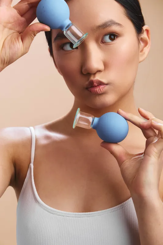 Asian woman uses facial cupping tools in a calm home setting, applying gentle suction to her cheeks with a focused expression, showcasing a modern skincare routine.
