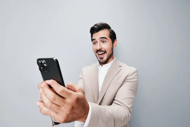 Улыбающийся мужчина в сером деловом костюме держит смартфон, стоя в студии на бежевом фоне