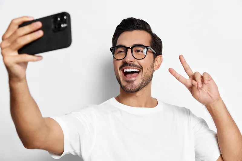 Happy smiling young man wearing glasses taking a selfie with a smartphone while showing peace sign on a solid light background. People lifestyle concept.