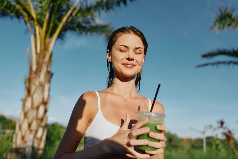 Smiling woman in a white tank holds a green smoothie outdoors under bright sun, radiating wellness and vitality. Outdoor lifestyle moment with tropical trees and blue sky.