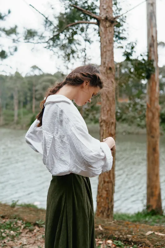 Young woman in a white shirt and green skirt standing by a tranquil lake, embracing nature with a contemplative expression and relaxed charm