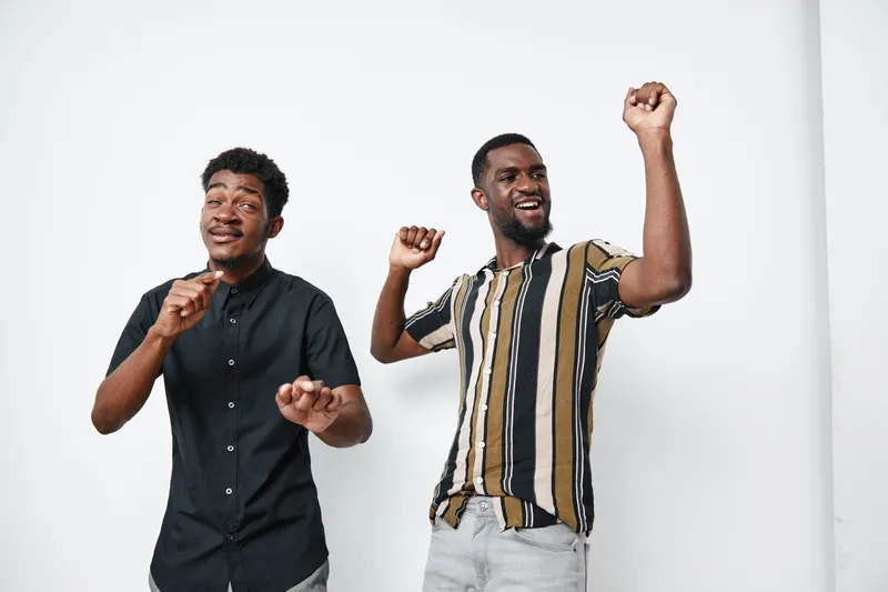Two young black men dancing and smiling with joy on white background. Casual clothing, happy mood, friendship and celebration with dark skin male friends together.