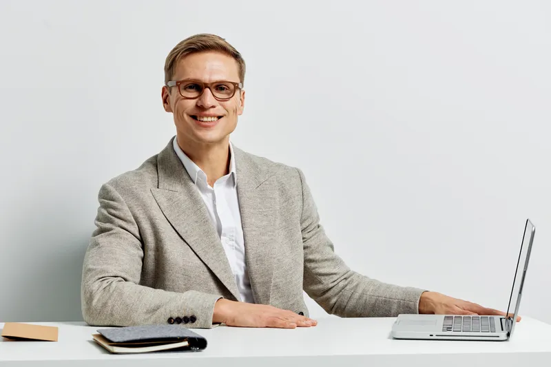 Professional middle-aged man in modern office wearing grey blazer and white shirt working on laptop smiling at camera against plain white background. business concept, office work, corporate lifestyle