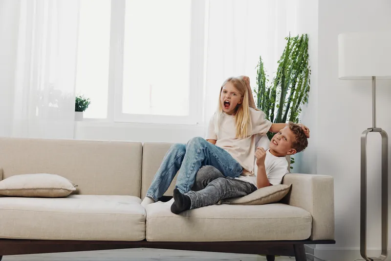 Two children playfully fight on a beige sofa in a bright living room with large windows and green plants. The scene captures joyful childhood energy and sibling interaction.