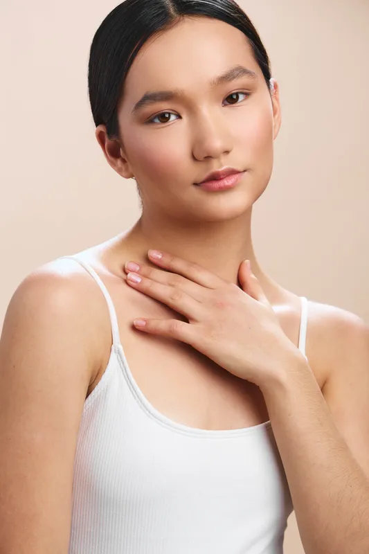 asian woman poses in a plain studio, wearing a white tank top, with a gentle smile and hand resting on her chest, conveying calm confidence and poised simplicity.