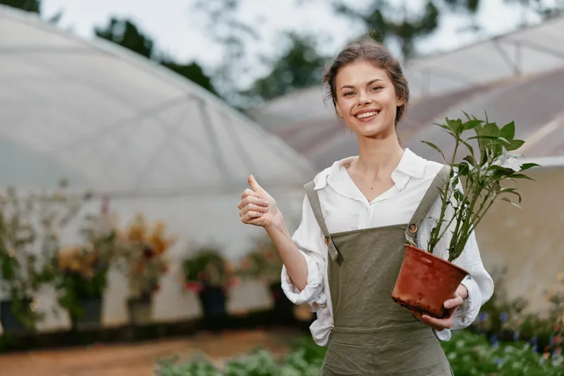 Young woman in overalls joyfully holding a potted plant with a thumbs up gesture in a botanical garden setting, reflecting a vibrant, healthy lifestyle