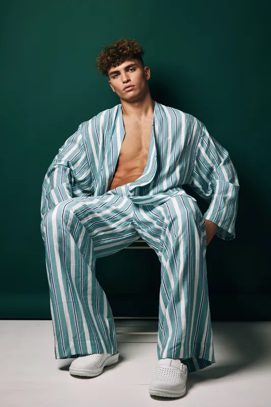 Young man with curly hair wearing striped loose pajamas and white slippers sitting on chair against dark green background in studio. Casual fashion and relaxed pose.