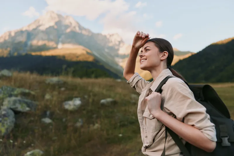 Young woman with a backpack hikes through a mountain meadow, shielding eyes from the sun, smiling toward distant peaks and a clear blue sky, her khaki shirt and rugged pants ready for the trail