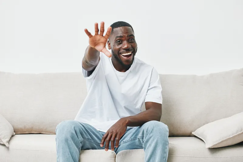 Smiling African American man greeting with a hand wave, sitting relaxed on a light sofa, wearing a casual white t shirt and jeans, creating a welcoming atmosphere