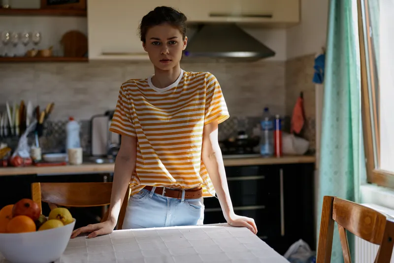 Casual woman stands in a bright kitchen leaning on a table, wearing a striped tshirt and jeans, everyday domestic scene with natural light