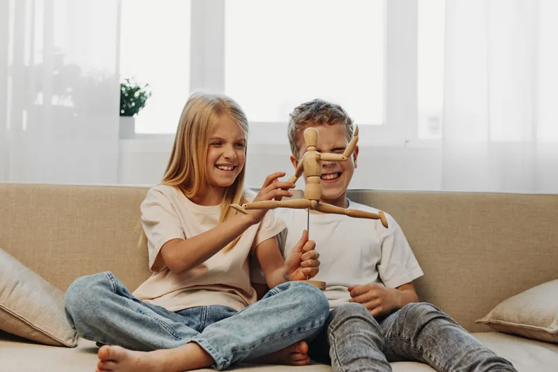 Happy children playing with wooden puppet on sofa in bright living room, smiling boy and girl enjoying creative fun together.
